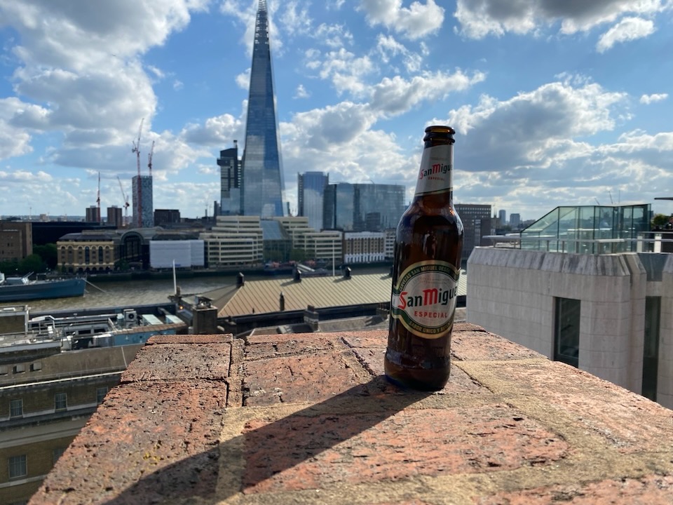 A bottle of beer sat on a little wall with a view of London, the Shard and a blue sky in the background