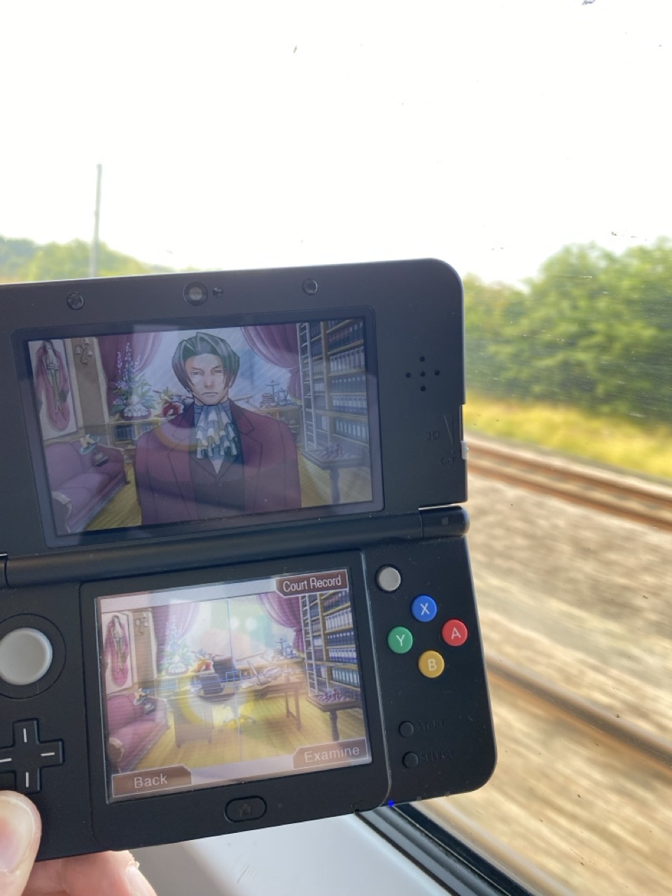 A 3DS running Phoenix Wright, with the view out a train window behind it.