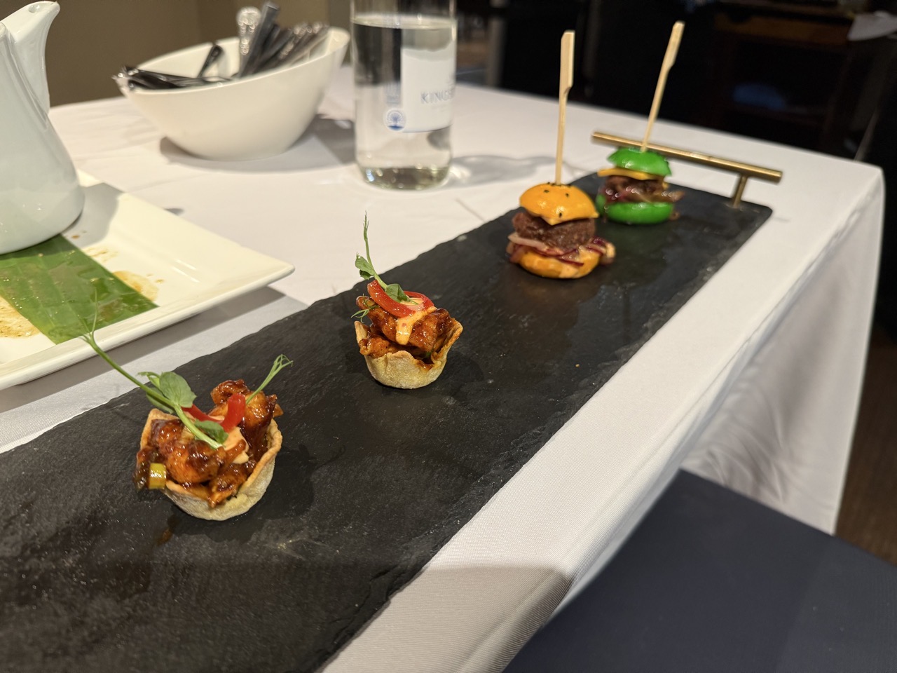 Some little canapés on a slate. The two nearest the camera are little taco looking things, the others are lamb in brightly coloured tiny bread rolls